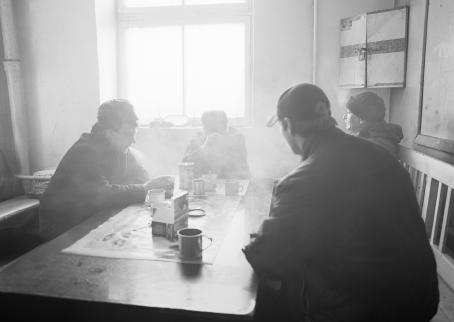 Mustavalkoisessa kuvassa miehiä istuu pöydän ääressä kahvilla. Ikkunasta tuleva valo on kirkasta ja tekee kuvaan utuisen tunnelman.