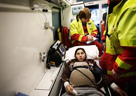 Raskaana oleva romaninainen makaa ambulanssin sängyllä. Hänen ympärillään on kaksi keltapunaisiin asuihin pukeutunutta ensihoitajaa. 