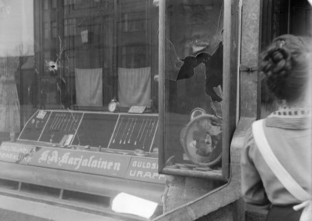 Mustavalkoisessa kuvassa liikkeen näyteikkuna, josta lasi on mennyt osittain rikki. Ikkunassa on esillä koruja, kelloja ja valokuvia. Ikkunan alla lukee "Kulta ja kellosepänliike K.A. Karjalainen".