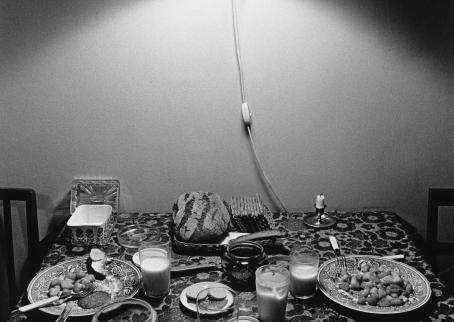 Mustavalkoinen kuva ruokapöydästä, jossa on ateria kesken. Pöydän päällä on lamppu joka valaisee pöydän.