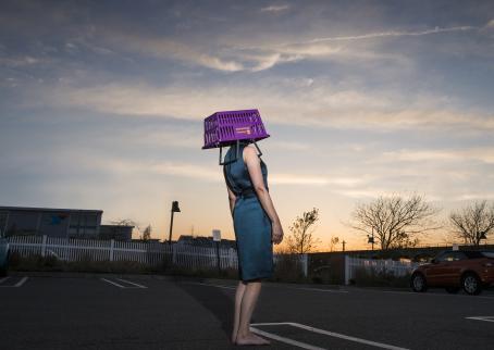 Nainen sinisessä mekossa seisoo keskellä asfalttista parkkipaikkaa violetti ostoskori päässään. Kuva on otettu alaviistosta, ja naisen takana näkyy auringonlaskun hieman oranssiksi värjäämä taivas.