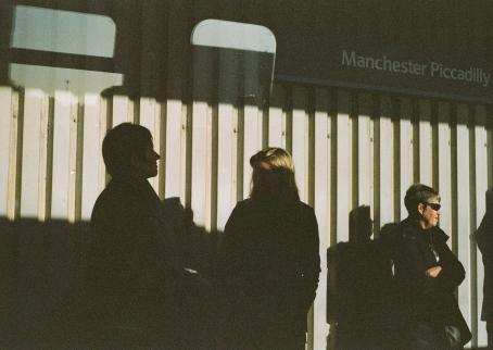 Ihmisiä seisoo seinän edessä. Osa ihmisistä jää varjoon, osa on auringossa. Ylhäällä lukee Manchester Piccadilly.