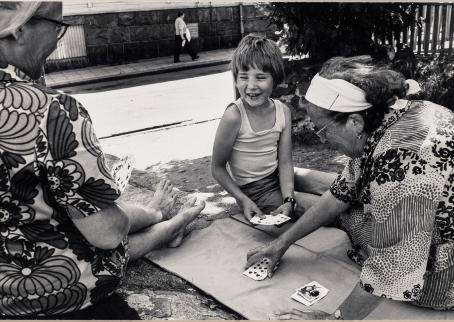 Mustavalkoisessa kuvassa kaksi vanhempaa naista ja lapsi pelaavat korttia puistossa maassa olevan alustan päällä. Aurinko paistaa. Lapsi hymyilee. 