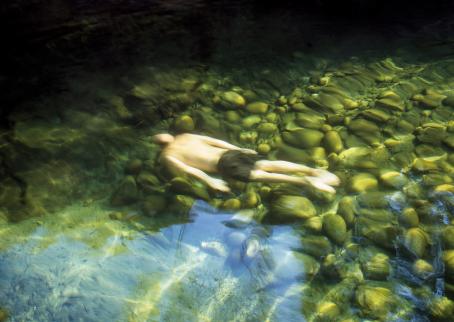 Veden alla makaa mies kasvot pohjaan päin, pelkät mustat shortsit jalassa. Pohja on kivinen ja vihreän levän peitossa. 
