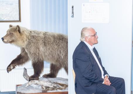 Vanhempi mies sinisessä puvussa istuu tuolilla metallisen oven edessä. Hänen takanaan seinän vieressä on täytetty karhu, joka katsoo eri suuntaan kuin mies.