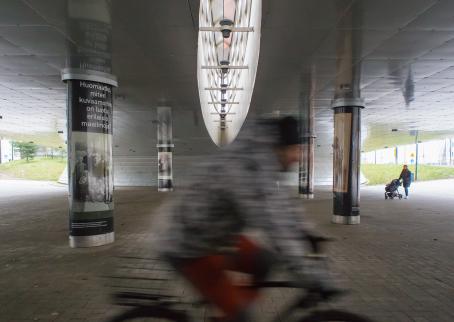  A view of a pedestrian and cycling path under a bridge. In the foreground is a blurry cyclist. In one pillar of the bridge there is an advertisement that has an old black and and white photograph. The ad says: " Do you notice how different worlds have been created in studios?"