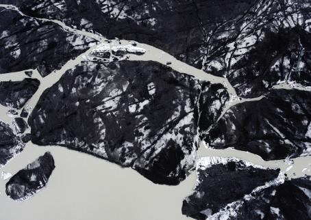 Korkealta ylhäältä päin otettu kuva, jossa valkoinen jäätikkö on lähes täysin mustana hiilipölystä. Keskellä menee vaaleita juovia, kuin jokia halkoen jäätikköä.