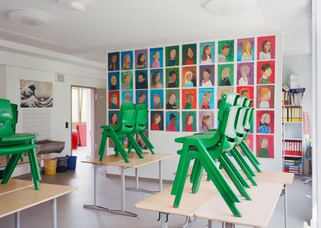 Luokkahuonemaisessa tilassa pöydille on nostettu vihreitä tuoleja kasoihin. Yksi seinä on täynnä piirrettyjä muotokuvia lapsista. Yhdellä seinällä on allas jonka alla on roskakoreja. Tilasta johtaa ovi myös toiseen huoneeseen ja käytävään. 