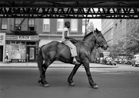 A black and white picture, where a black man is riding a big, dark horse on a city street. The horse is not wearing a saddle, only a bridle.