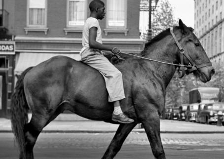 A black and white picture, where a black man is riding a big, dark horse on a city street. The horse is not wearing a saddle, only a bridle.