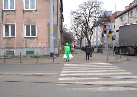 A woman in a green full body skinsuit that covers her face too, stands in front of a crosswalk at a red light. She is carrying four white shopping bags. Next to her is a woman in a brown jacket looking at her. 