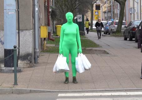 A woman in a green full body skinsuit that covers her face too, stands in front of a crosswalk at a red light. She is carrying four white shopping bags. Next to her is a woman in a brown jacket looking at her. 