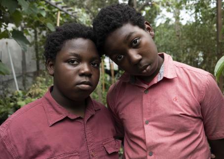 Two black children. Other is a bit taller, and is leaning on the other’s head. Both have a light red collared shirt and both are looking straight into the camera. 