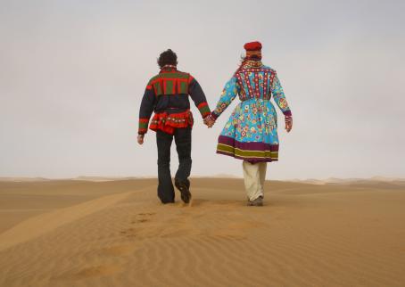 Kaksi henkilöä kävelee käsi kädessä hiekalla aavikolla. Heillä on päällään värikkäät vaatteet.