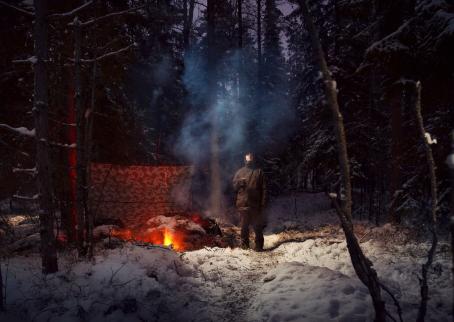 Mies seisoo lumisessa metsässä muki kädessään ja katsoo ylöspäin. Hänen vieressään palaa nuotio. Nuotion vieressä on puiden väliin pingotettu kangas.