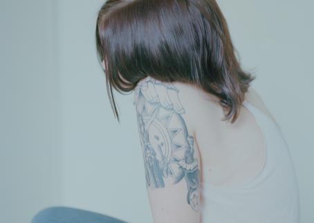 Nuori henkilö istuu sivuttain, hiukset peittävät kasvot. Hänellä on valkoinen toppi ja olkavarressa tatuointi.
