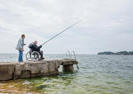 Vanha mies pyörätuolissa on onkimassa kivisellä laiturilla. Hänen vieressään on vanha nainen.