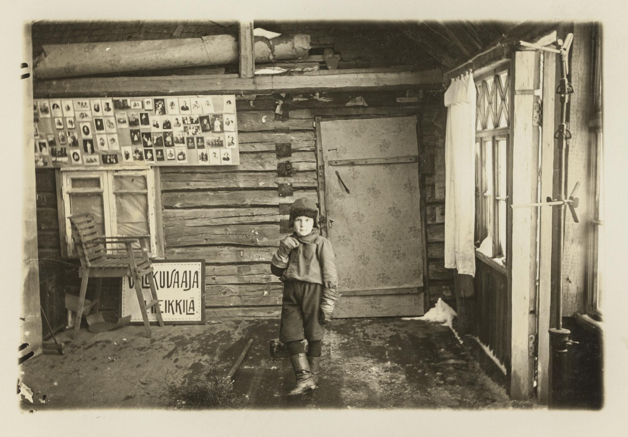 Ilmari Heikkilän studio Raivolassa, 1921. D1974:33/13332, Suomen valokuvataiteen museon kokoelma. Mustavalkoisessa kuvassa lapsi seisoo kuvan keskellä keskellä hirsitalossa myssy päässään ja nahkasaappaat jaloissaan.