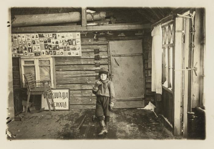  Mustavalkoisessa kuvassa lapsi seisoo kuvan keskellä keskellä hirsitalossa myssy päässään ja nahkasaappaat jaloissaan.
