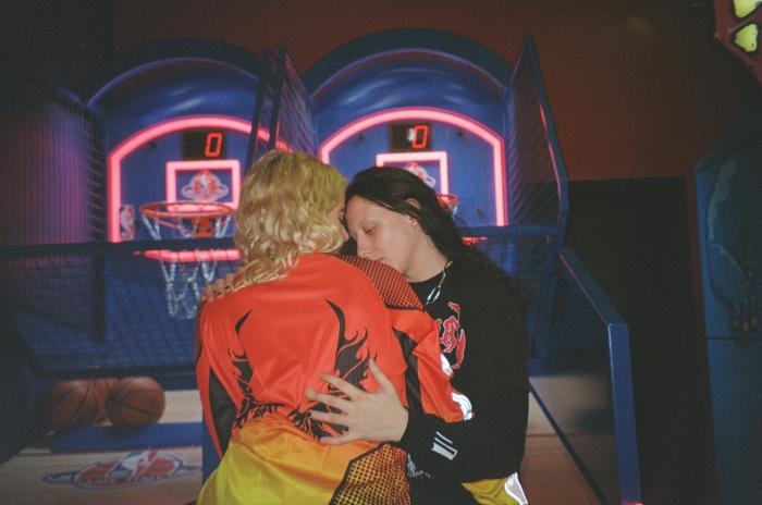 A dark‑clothed, black‑haired person and a colorfully dressed, blond‑haired person are hugging. In the background, there is an arcade basketball game.