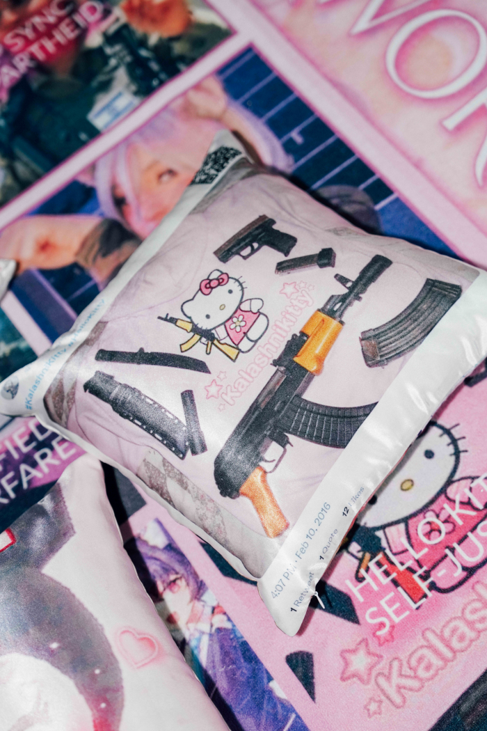 Tyyny, jossa rynnäkkökivääriä pitelevä Hello Kitty -hahmo. Hahmon ympärillä erilaisia aseita. Tyynyn taustalla videopelihahmo ja vaaleanpunaista söpöä estetiikkaa.