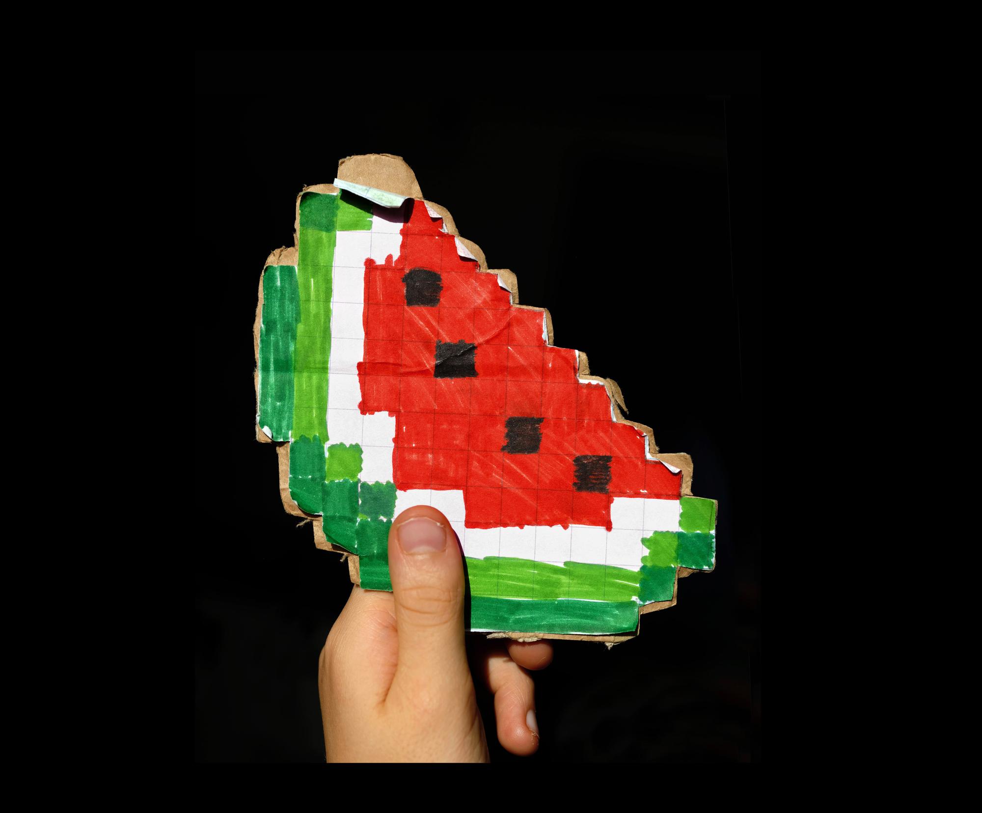 Käsi pitelee paperista ja pahvista tehtyä vesimelonin kuvaa mustaa taustaa vasten. Vesimeloni koostuu suurista pikseleistä.